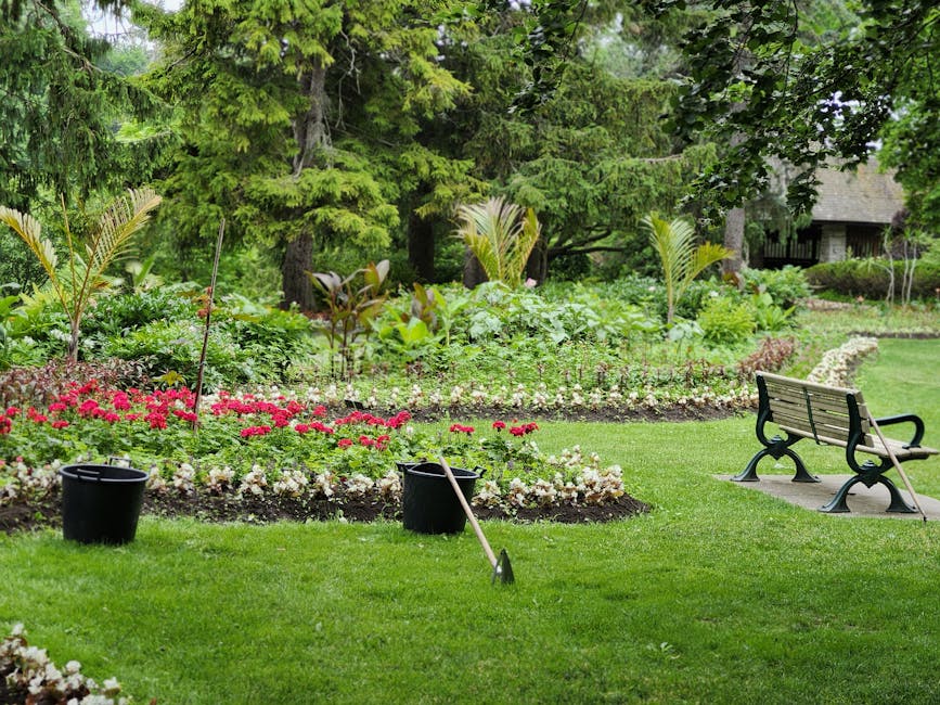 A lush, well-maintained garden featuring a vibrant, green lawn with freshly trimmed grass. In the foreground, two black buckets and a garden hoe are positioned on the grass, indicating ongoing garden 