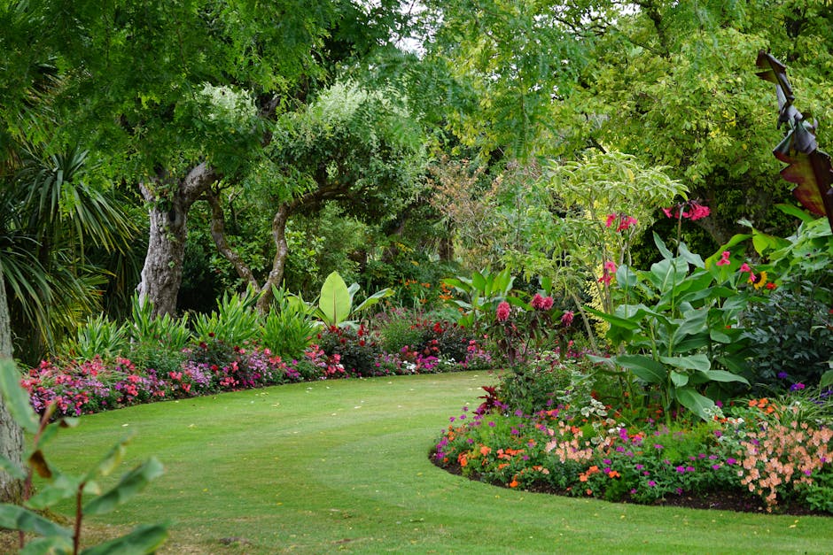 A lush, well-maintained garden featuring a vibrant, green lawn with freshly trimmed grass. In the foreground, two black buckets and a garden hoe are positioned on the grass, indicating ongoing garden 