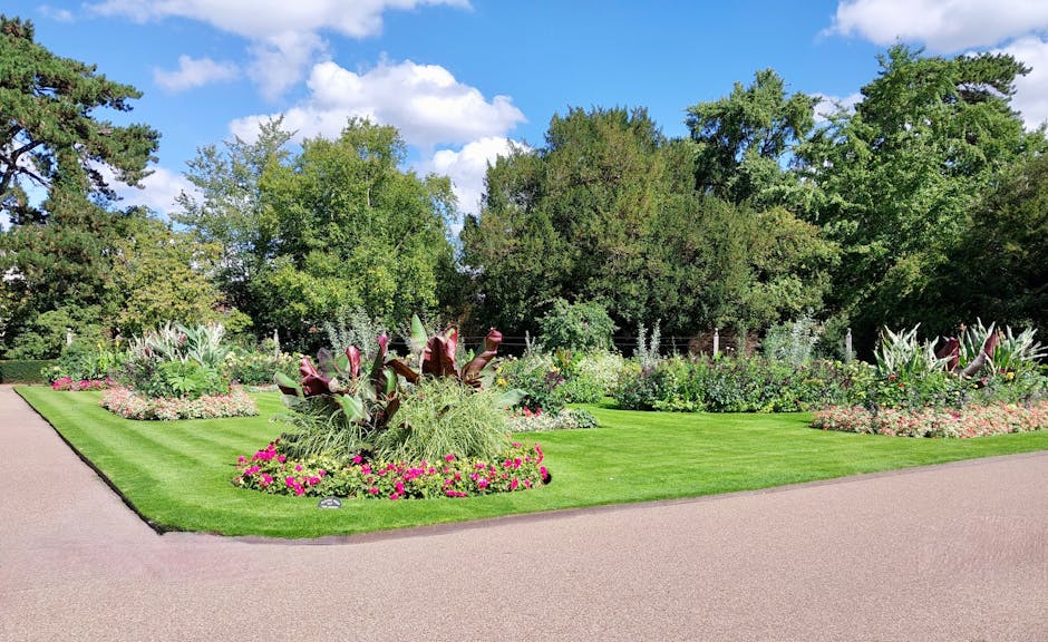 A neatly landscaped garden featuring a well-maintained, lush green lawn bordered by garden beds filled with a variety of flowering plants and ornamental foliage. The beds contain vibrant pink, purple,