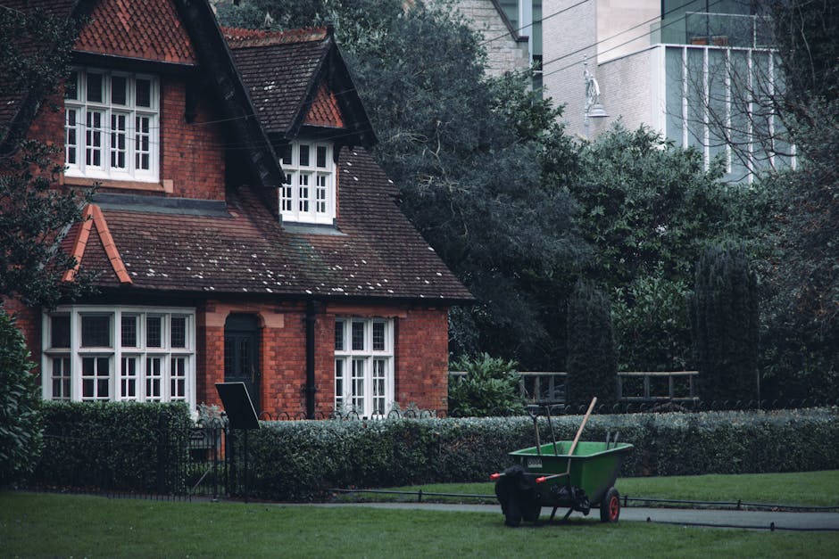 A residential garden scene featuring a well-maintained lawn bordered by neatly trimmed hedges and a low decorative metal fence. In the foreground, a green wheelbarrow with a black handle and red wheel